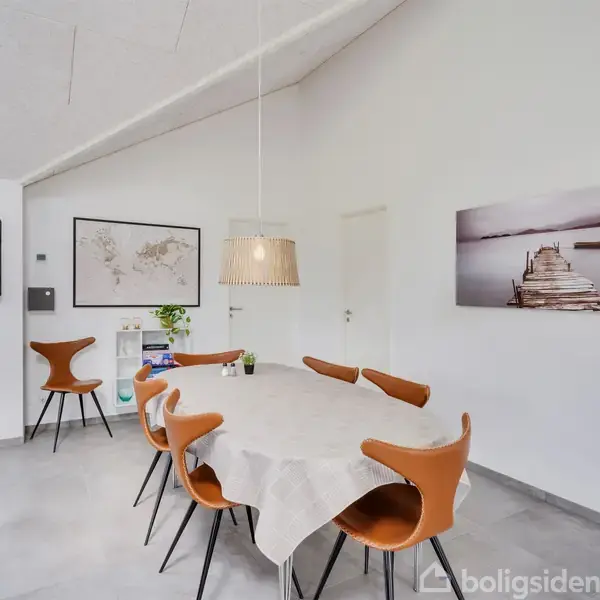 Et spisebord dækket med en dug omgivet af seks moderne, brune stole. En hængelampe er tændt over bordet. Væggen har et verdenskort og et billede af en bro.