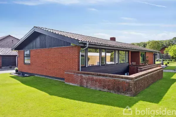 Rødt murstenshus står stille i en grøn have med velplejet græsplæne. Bygningen har store vinduer og en veranda indrammet af en murstensmur. Solrig himmel i baggrunden.