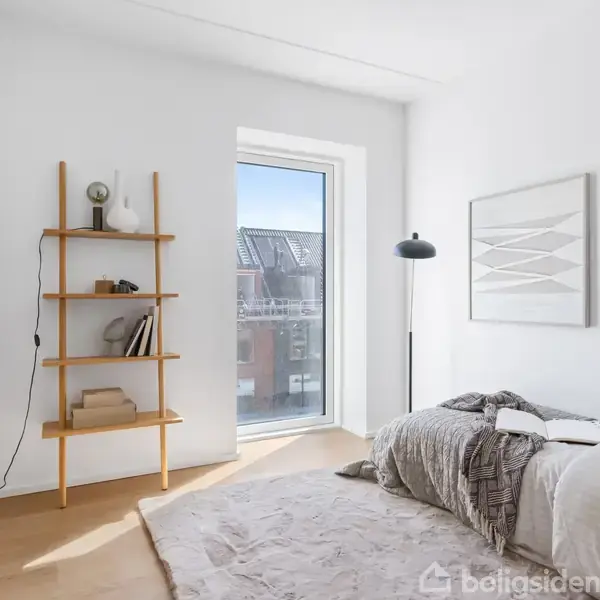Et lyst værelse har en enkel seng med gråt sengetæppe. En træhylde ved siden af indeholder bøger og en lampe. Store vinduer giver udsigt til bygninger udenfor.