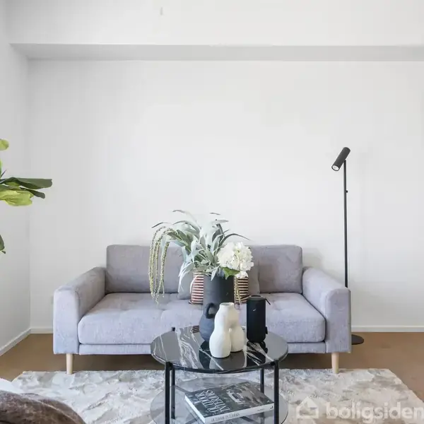 Sofa med puder og blomsterdekorationer står i en lys stue. Et stuebord med vaser foran, lampe til højre, plante til venstre og et spejl på væggen.