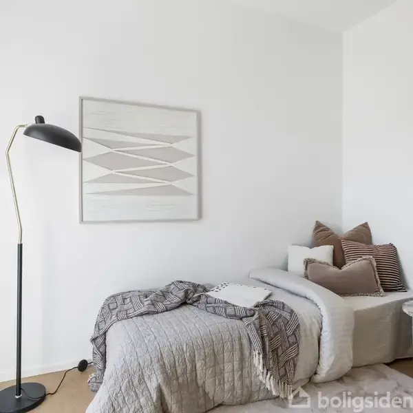 En enkelt seng er redt med gråt sengetøj og pyntet med brune puder. Ved siden af står en sort gulvlampe. Væggene er hvide, og der hænger et geometrisk billede. En grøn plante står på en lille bord.