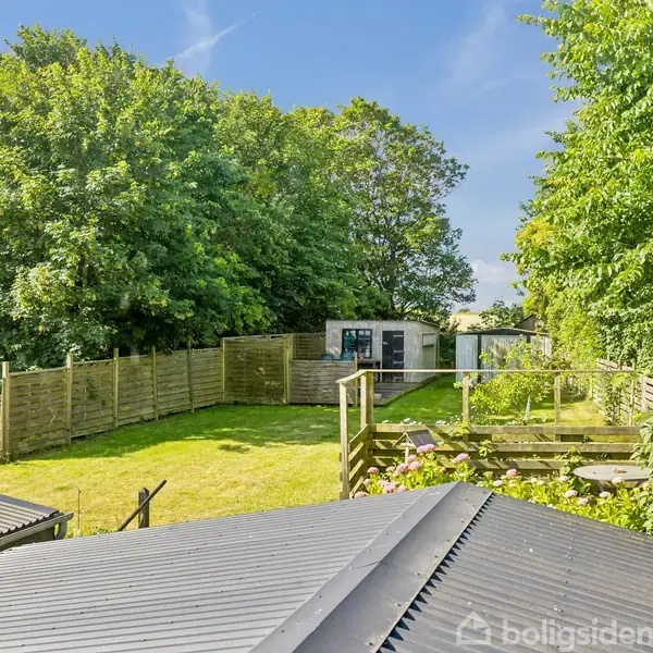 En have med grøn græsplæne omgivet af træer og hegn, hvor et skur står bagerst. Tagene af bygninger kan ses forrest i billedet.