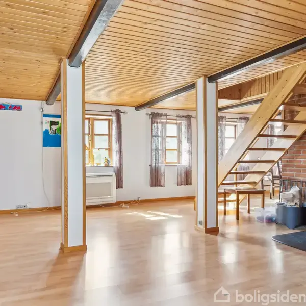 Stue med brændeovn tændt, trappe til første sal, trægulve og loft. Væggene har kunstværker, og der er flere vinduer med gardiner, der lader dagslys ind.