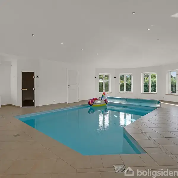 En lys indendørs pool fyldt med vand, omkranset af beige fliser. Store vinduer bagved lader dagslys komme ind. En farverig poolflyder flyder i vandet, og en stol står til højre.