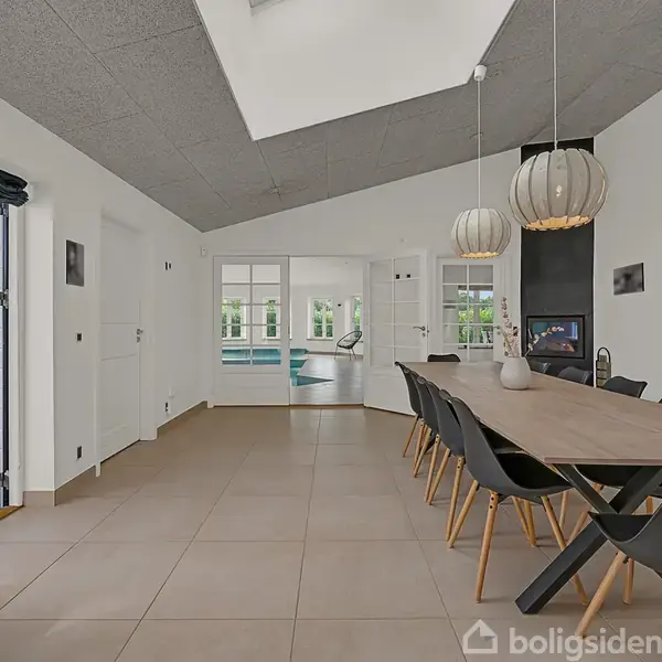 Et moderne spisebord med otte sorte stole står i et rummeligt, lyst køkken-alrum. Over bordet hænger tre dekorative lamper. I baggrunden ses en døråbning til et rum med pool.