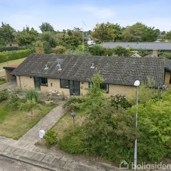 Luftfoto af en landsby med en hvid pil, der peger på et bestemt hus. Omgivet af grønne marker og træer. Flere veje og boliger kan ses i området.
