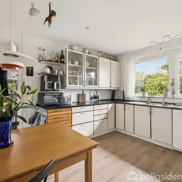 Et lyst køkken med hvide skabe og sorte bordplader. En blå vase med blomster står på et træbord. Store vinduer viser en grøn have udenfor.
