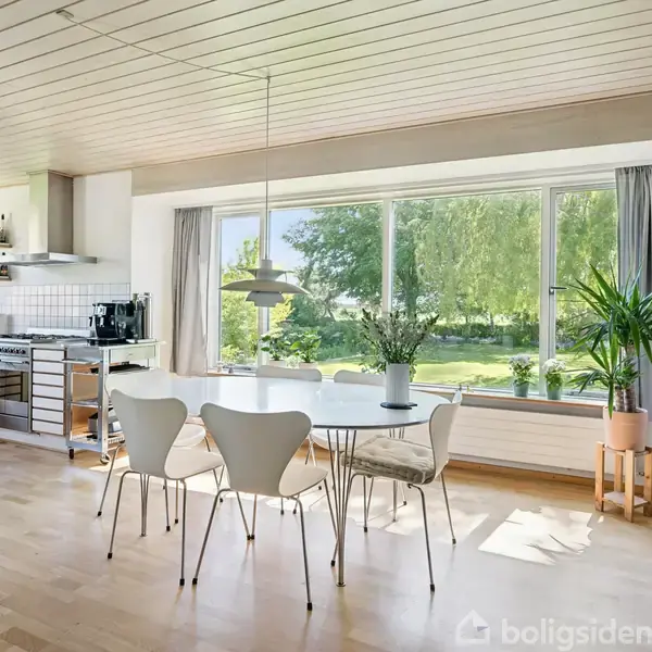 Et moderne køkkenalrum med hvide møbler og en rund spisebord med fire stole. Store vinduer giver udsigt til en frodig have med træer, og rummet er fyldt med naturligt lys.