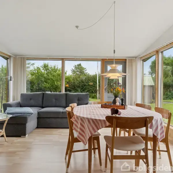 Gråt L-formet sofa står i stue med store vinduer omkring. Rundt bord med ternet dug og fire stole i forgrunden. Udsigt til grøn have gennem vinduerne.