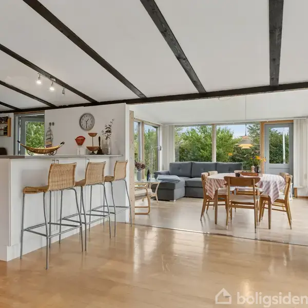 Køkken-alrum med høje stole ved en bar og spisebord, der står i forgrunden. Lysesofa og store vinduer giver udsigt til grønne træer udenfor.