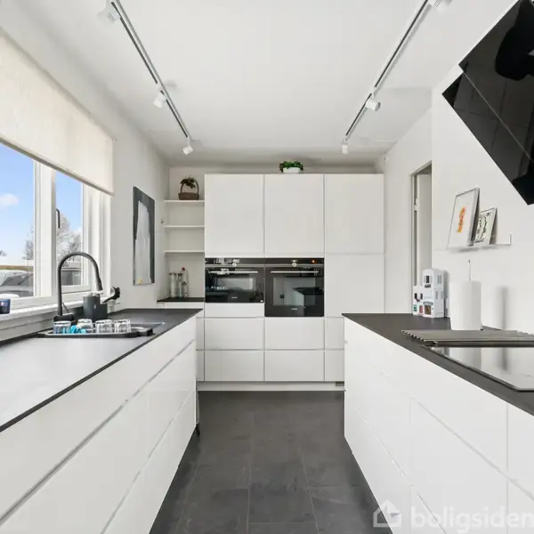 Et moderne køkken med hvide skabe og sorte bordplader, hvor der ses en indbygget ovn. En kaffemaskine står på bordet ved vinduet, der slipper dagslys ind.