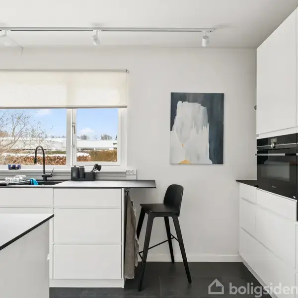 Køkkenet har hvide skabe og sorte bordplader. En sort stol står ved køkkenøen. Store vinduer giver udsigt til en sneklædt have. Maleri hænger på væggen.