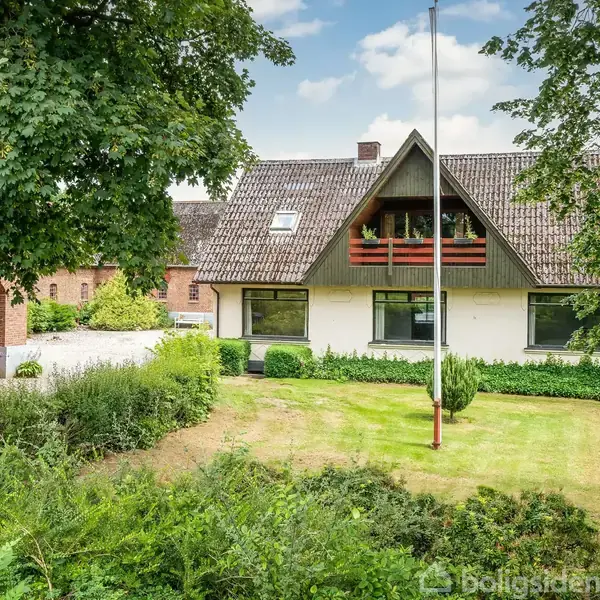 Hus med græsplæne og træer, stående i en frodig have. Det har en rød garage og hævet balkon. Blå himmel og et flagstang eksisterer i omgivelserne.
