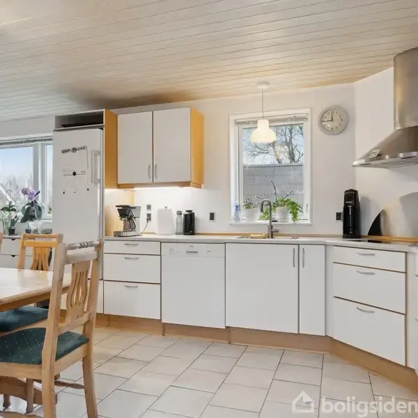Køkken med hvide skabe, stålkøkkenapparater og en rund spisebord med stole. To vinduer bringer lys ind og blomster tilføjer dekoration. Flisegulv og en loftslampe skaber en hyggelig atmosfære.