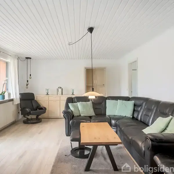 En stue med sort lædersofa, grønne puder, og en træsofabord. Der er en sort lænestol, et skab i baggrunden og store vinduer med hvide gardiner.
