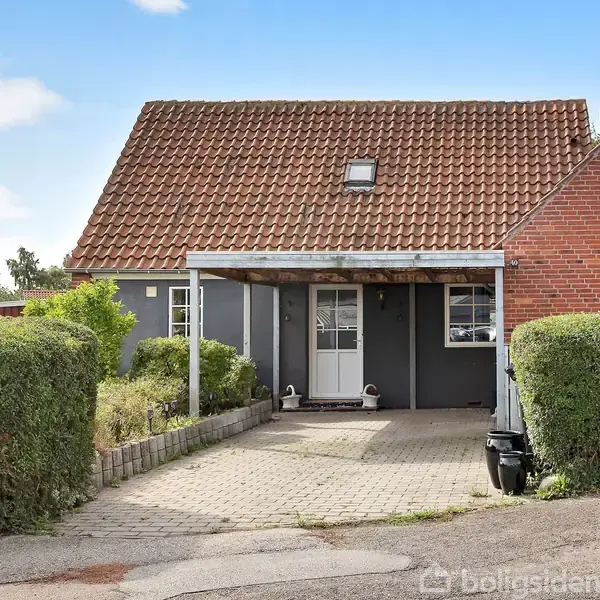 Et hus med rødt tegltag står stille i en boligkvarter, omgivet af en klippet hæk og en indkørsel af fliser. Til venstre ses et skilt med "TIL SALG" og et telefonnummer.