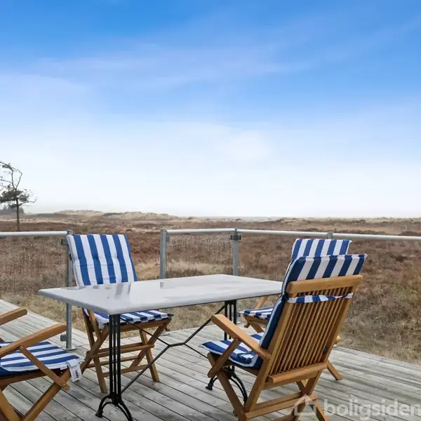 Terrassemøbler med blå-hvidstribede hynder står på en træterrasse. Baggrunden viser en vid udsigt over et øde klitlandskab under en klar blå himmel.
