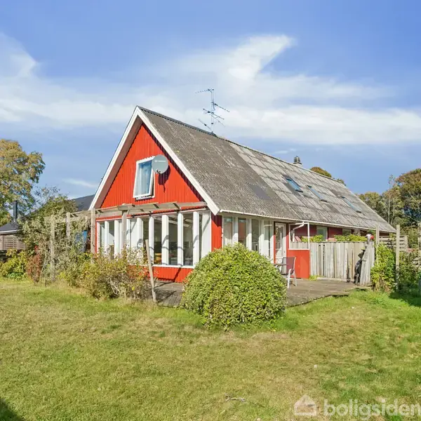 Rødt træhus med skråt tag står i en frodig have med buske og græsplæne. Sollys oplyser huset, der har et stort vindue på facaden.