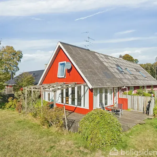 Rødt hus med skråt tag ses fra haven, omkranset af grønne buske og træer. Solrige omgivelser under en klar blå himmel. En terrasse med havemøbler er synlig.