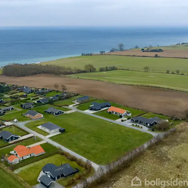Huse med orange og sorte tage står stille på grønne grunde, omgivet af marker og nær en kystlinje med udsigt over havet under en overskyet himmel.