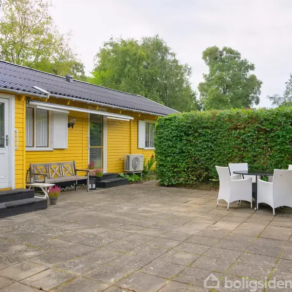 Gulmalet hus står ved siden af flisebelagt terrasse, hvor et rundt bord med hvide stole er placeret. En høj hæk danner baggrund, omgivet af grønne træer.