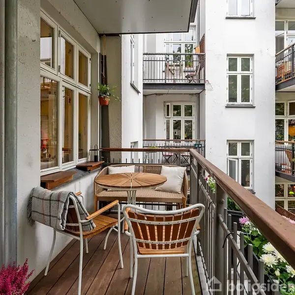 Et balkonbord og stole står på en træterrasse. Tæpper ligger på stolene ved siden af blomster. Omgivet af en hvid bygning med flere altaner og grønne planter tæt på.