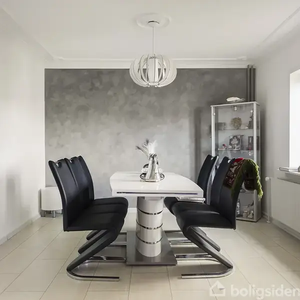 Et spisebord med sortpolstrede stole står i et lyst rum. En moderne lampe hænger over bordet, og en vitrineskab ses ved siden af et vindue.