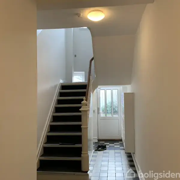 Trappe op til øverste etage, beklædt med sort tæppe og hvidt gelænder. Ved siden af trappen en entre med flisegulv og en hoveddør i lys, spartansk indretning.