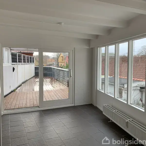 En lys stue med store vinduer og flisegulv har udsigt til en udendørs træterrasse. Der er radiatorer under vinduerne, og tagene på nærliggende bygninger er synlige udenfor.