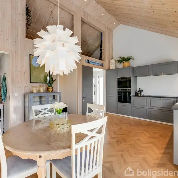 Et rundt spisebord med fire stole står centralt i et lyst køkkenalrum. Over bordet hænger en moderne, hvid lampe. Køkkenet har grå skabe og ligger langs væggen.