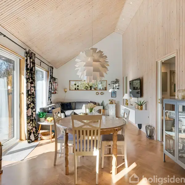 Et lyst rum med spisebord og stole i træ, under en stor, hvid lampeskærm i moderne stil. Udgang til terrasse gennem glasskydedøre omgivet af grønne planter og sofaområde.