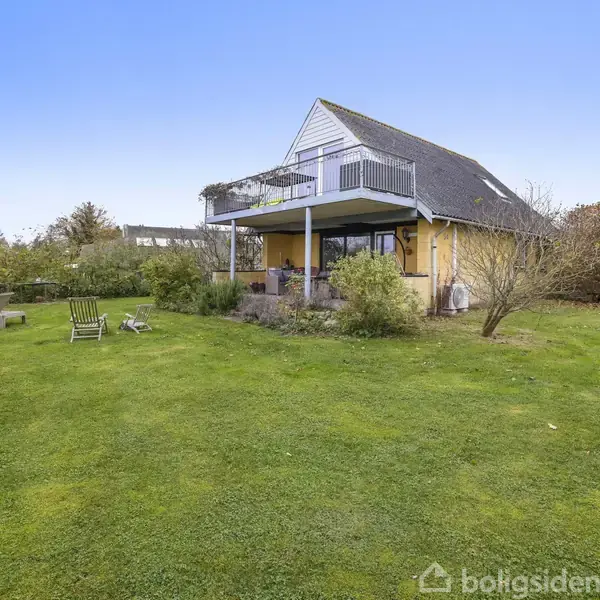 Et gult hus med sort tag står i en velplejet have med en terrasse og balkon. I haven er der et lille haveskur og havemøbler omgivet af buske og træer.