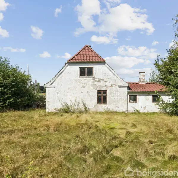 Et hvidt hus med rødt tegltag står stille omgivet af højt græs og buske under en blå himmel med lette skyer.