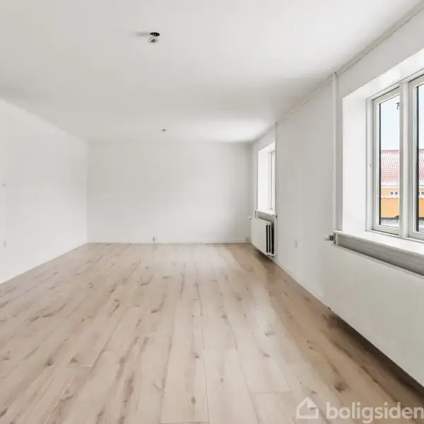 Rummelig, lys stue med trægulv og hvide vægge. To store vinduer lader naturligt lys ind. Der er en synlig radiator under vinduerne. Moderne og indbydende atmosfære.