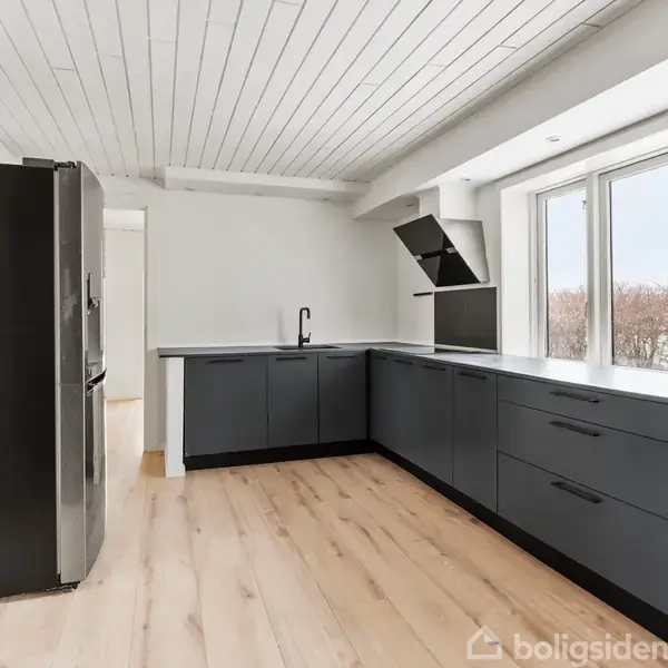 Køkken med mørkegrå skabe, moderne apparater og en stor køleskab. Store vinduer slipper naturligt lys ind. Trægulv og hvid loftbeklædning skaber en lys og rummelig atmosfære.
