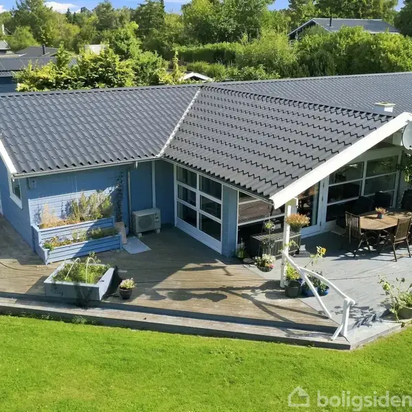 Et blåmalet hus med sort tag står på en træterrasse. Ved siden af huset er der havemøbler og planter. Huset er omgivet af en grøn græsplæne og træer.