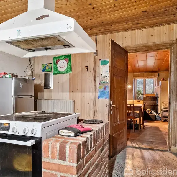 Komfur står stille i et køkken med træbeklædning, omgivet af en murstensbarriere. I baggrunden ses en åbentstående dør til en spisestue med et bord og stole. En tegning hænger på væggen.