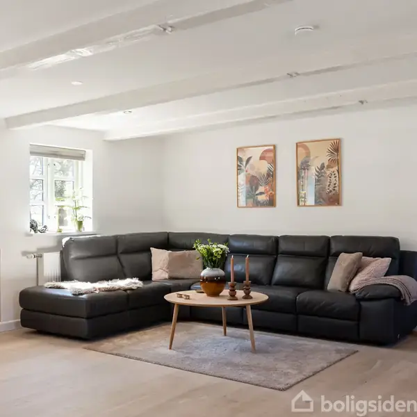 Sort læderhjørnesofa står i et lyst stueområde med trægulv. Et rundt sofabord er pyntet med dekorationer. Væggene er dekoreret med to kunstbilleder. En skænk og en lampe er til venstre.