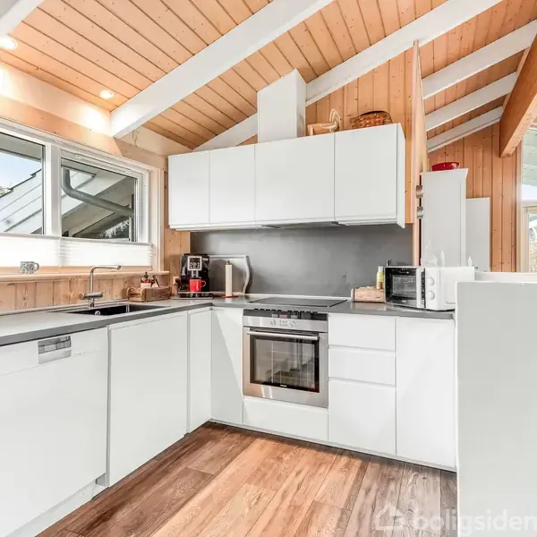 Et moderne køkken med hvide skabe og apparater. En ovn og opvaskemaskine er indbygget. Køkkenet har et skråt træloft og store vinduer giver naturligt lys.