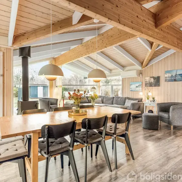 Spisestue med et stort træbord, omgivet af stole i et rum med synlige træbjælker. Store vinduer lukker lys ind, og der er udsigt til en have. Pejs ved siden af bordet.