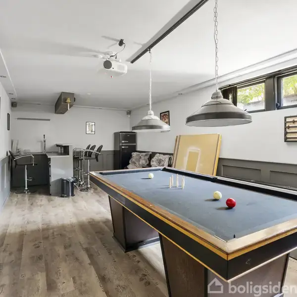 Terrasse med havemøbler arrangeret omkring et spisebord dækket med ternede duge. Sofa og stole står i højre side, omgivet af en træhegn og grønne træer i baggrunden.