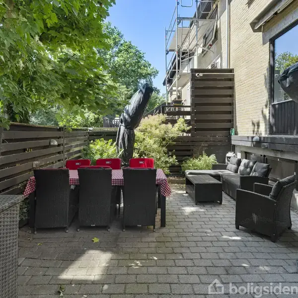 En udendørs terrasse med et langt bord dækket af rød-hvidternede duge, omgivet af sorte kurvestole. Sofaer og en parasol findes også, med grønne træer rundt om.