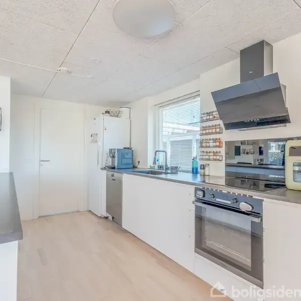 Et moderne køkken med hvide skabe og grå bordplader, hvor en ovn og kogeplade er integreret. Rummet er lyst med vindue og dekoreret med blomster på venstre side.