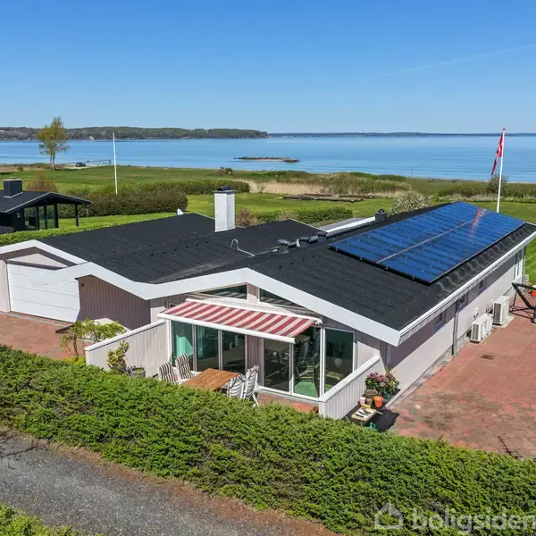 Hus med solpaneler på taget står stille omgivet af velplejet græsplæne. Solbeskinnet terrasse med markise i forgrund. Udsigt over blå sø og grønne omgivelser i baggrund.