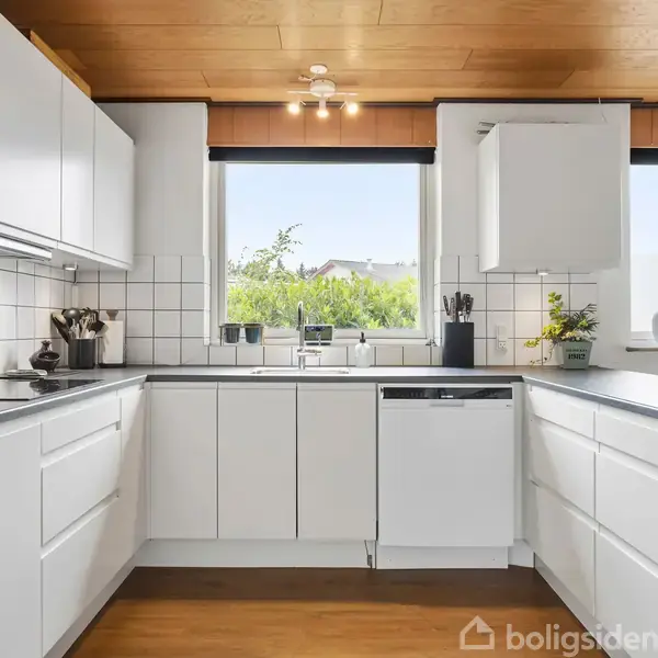 Hvidt køkken med moderne skabe omkranser en opvaskemaskine. Et vindue viser en udsigt til buske og træer, mens planter og køkkenredskaber pynter på den mørke bordplade.