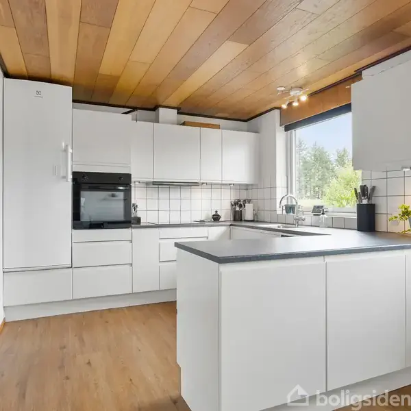 Køkken med hvide skabe og grå bordplader. Der er indbyggede ovne, en vask, og elementer som planter og køkkenartikler. Træloft og vindue med udsigt til grønne omgivelser.