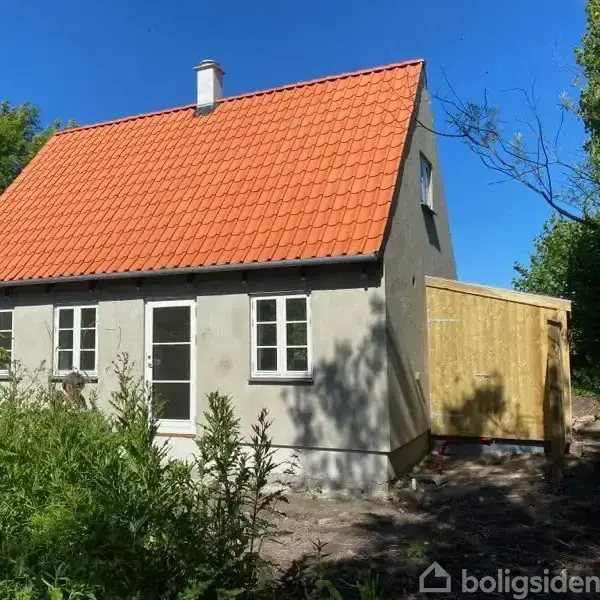 Et lille hus med rødt tegltag og grå facade står blandt høje buske. Huset har hvide vinduer og en tilstødende træskur. Blå himmel og træer omgiver stedet.
