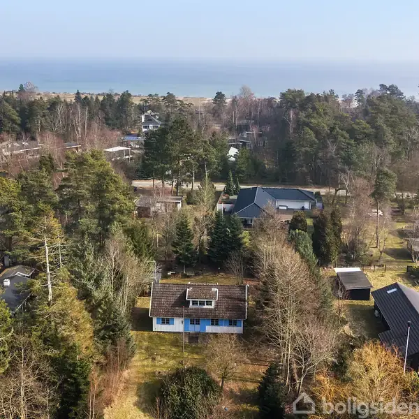Hus med blå vægge står i en skovfyldt boligområde nær kysten. Omkringliggende huse og træer skaber et tæt bebygget kvarter. Havudsigt i baggrunden under en klar himmel.