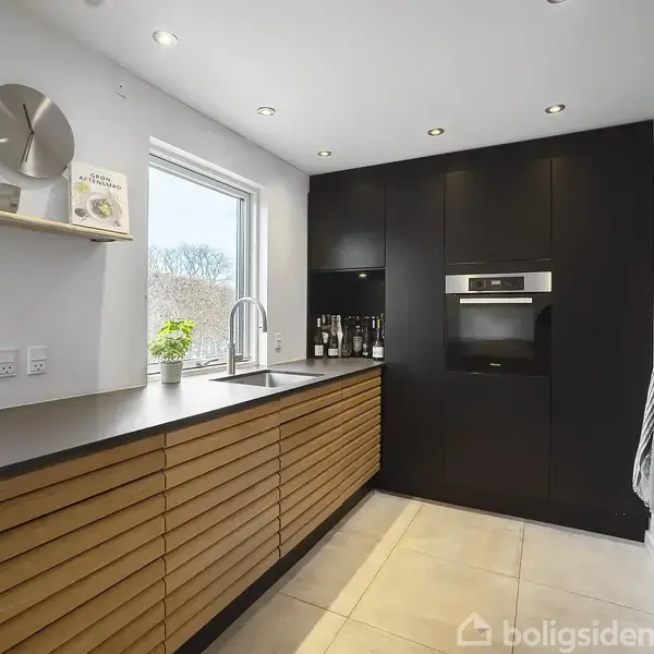 En moderne køkkenindretning med træskabe og sort køkkenudstyr. Et indbygget ovn er placeret i en sort væg. Et vindue og planter giver naturligt lys og liv.