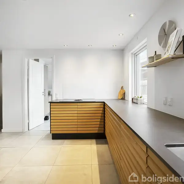 Køkken med træskabslåger og sorte bordplader. På væggen hænger en hylde med dekorative elementer, bl.a. en ur. Lyse fliser på gulvet og et stort vindue med planter.